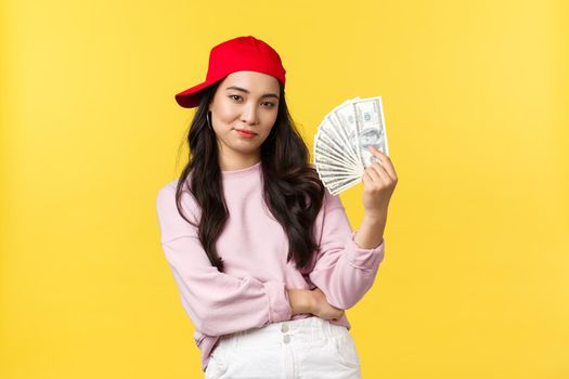 People emotions, lifestyle leisure and beauty concept. Sassy and confident, cool asian girl with lots of money, smiling determined, going shopping, ready to waste all cash, yellow background.