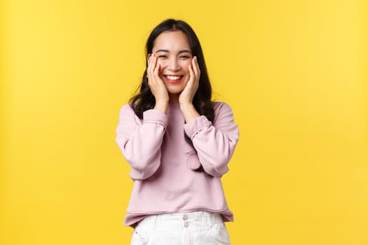 People emotions, lifestyle and fashion concept. Adorable smiling korean girl looking happy, rejoicing over big amazing news, hold hands on face and standing yellow background upbeat.