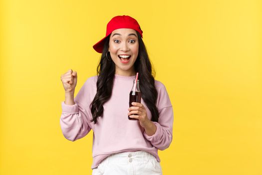 People emotions, drinks and summer leisure concept. Surprised excited cute asian girl in red cap, drinking soda beverage and chanting, watching sport game, enjoying vacation, yellow background.