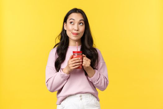 People emotions, lifestyle leisure and beauty concept. Dreamy and thoughtful, cute asian girl searching inspiration, looking up and thinking while drinking smoothie over yellow background.