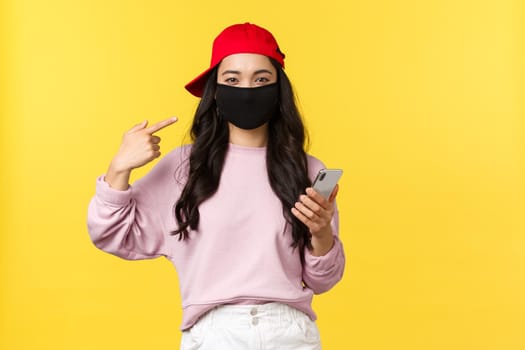 Covid-19, social-distancing lifestyle, prevent virus spread concept. Smiling cute asian girl pointing at face mask, asking protect health during coronavirus, holding mobile phone, yellow background.