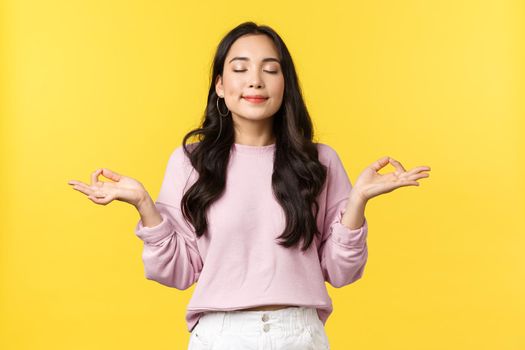 Lifestyle, emotions and advertisement concept. Patient and calm smiling beautiful asian girl keep body under control, inhale air, practice yoga breathing, meditating happy over yellow background.