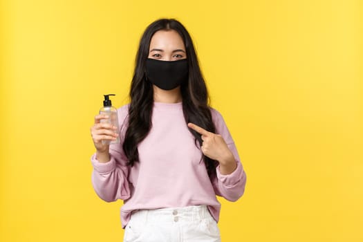 Covid-19, social-distancing lifestyle, prevent virus spread concept. Asian girl in face mask pointing at hand sanitizer, taking care personal hygiene during coronavirus pandemic, yellow background.