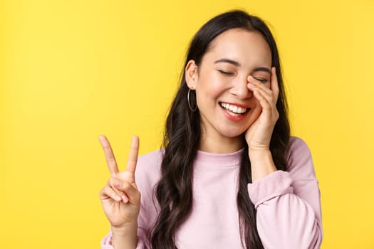 People emotions, lifestyle leisure and beauty concept. Close-up of carefree amused cute asian girl, showing peace sign touch face and laughing happy, standing yellow background.
