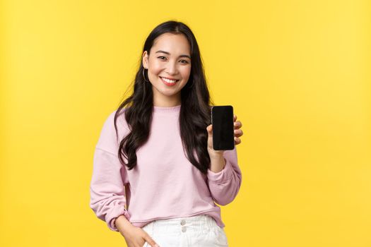 People emotions, lifestyle leisure and beauty concept. Smiling asian woman 20s, showing smartphone display, recommend application or mobile game, advertisement on screen, yellow background.
