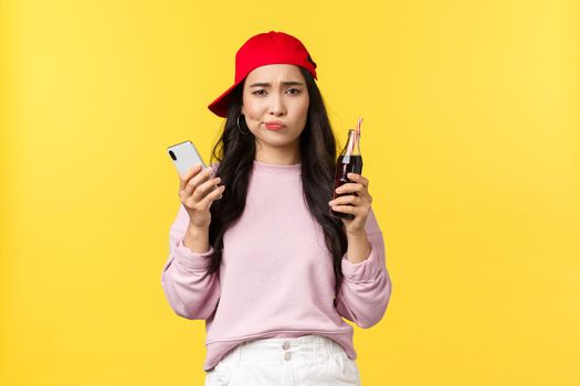 People emotions, drinks and summer leisure concept. Skeptical and displeased cute teenage asian girl in red cap, pouting disappointed as holding mobile phone and soda drink, yellow background.