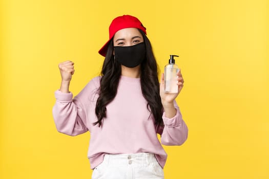 Covid-19, social-distancing lifestyle, prevent virus spread concept. Cheerful asian girl in face mask during coronavirus recommend hygiene product, showing hand sanitizer, yellow background.