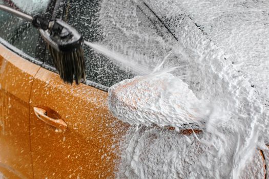 Detail on yellow car side mirror covered with thick white shampoo foam, more spraying, when washed in carwash.