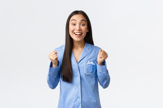 Hopeful excited asian girl in blue pajamas clench fists and looking rejoice camera, smiling as awaiting great news, feeling enthusiastic and upbeat as standing over white background.