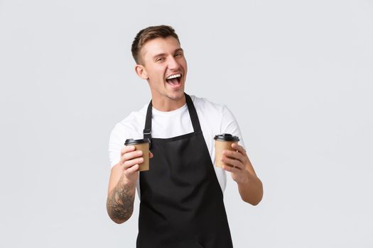Small business, coffee shop, cafe and restaurants concept. Enthusiastic happy barista in black apron holding two paper cups with coffee, selling and giving order to clients, laughing joyfully.