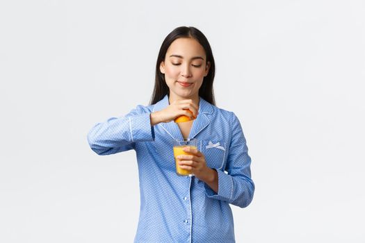 Morning, active and healthy lifestyle and home concept. Pretty healthy asian girl in pajamas prepare orange juice, squeezing orange in glass and smiling pleased, standing white background.