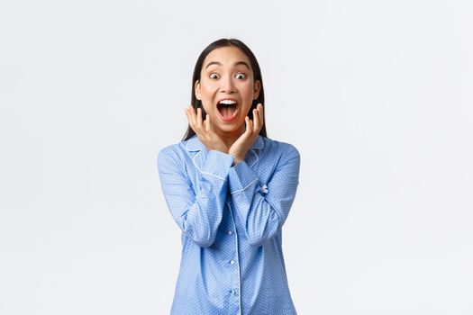 Excited super happy asian girl in blue pajamas waking up to awesome surprise, screaming from happiness looking at camera astounded, react to awesome gift, standing white background.