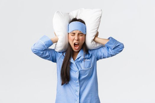 Angry furious asian girl in pajamas and sleeping mask, lying bed and shut ears with pillow, shouting mad as cant sleep from loud noise, neighbours through party at night, complaining annoying sound.