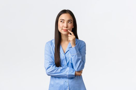 Cunning and thoughtful pretty asian girl in blue pajamas, smirk and looking upper left corner, imaging something, having interesting idea, standing white background in jammies.