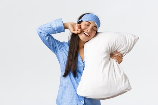 Smiling pleased cute asian girl in blue pyjama and sleeping mask, hugging pillow and stretching hands delighted as finally going bed, want sleep or waking up in morning, white background.