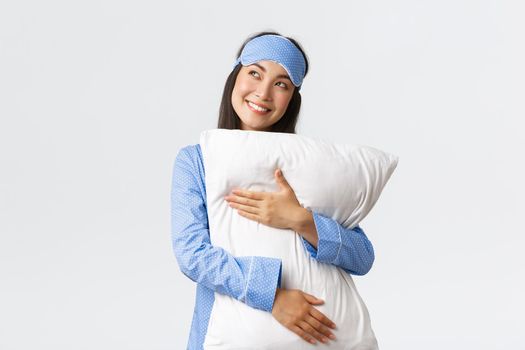 Dreamy beautiful asian girl in blue pajamas and sleeping mask hugging pillow and smiling pleased as looking upper left corner imaging something in bed, picturing, using imagination.