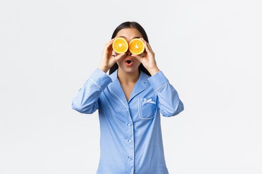 Morning, active and healthy lifestyle and home concept. Funny and playful cute asian girl in blue pajamas holding oranges over eyes, open mouth fascinated, look interested and impressed.