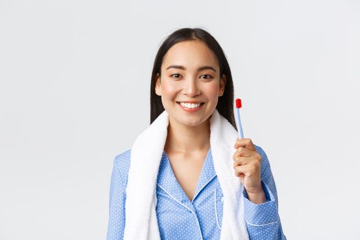 Daily routine, morning and hygiene concept. Pretty asian girl in blue pajama, holding towel and showing toothbrush, smiling white teeth, getting ready shower before bed, white background.