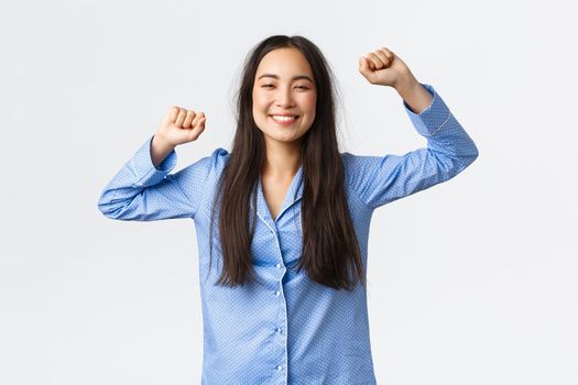 Pleased smiling korean girl in blue pajamas stretching delighted and grinning as waking up from good night sleep, have messy haircut from getting up bed, standing white background.