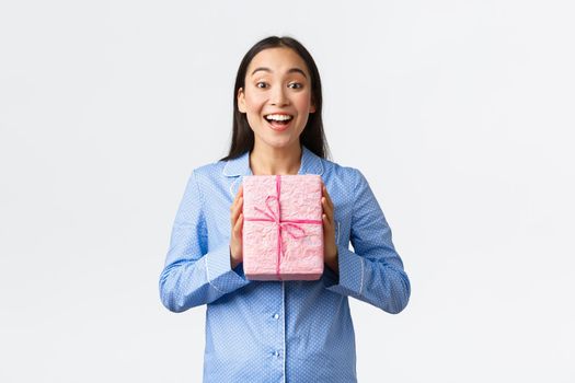Home, holidays and lifestyle concept. Happy and pleased smiling asian girl receive birthday present from family, holding gift as wearing pajama, unpack gifts on christmas eve, white background.