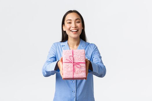 Home, holidays and lifestyle concept. Cheerful asian girl in blue pajama congratulate sister with christmas or birthday, giving gift and smiling, standing white background.
