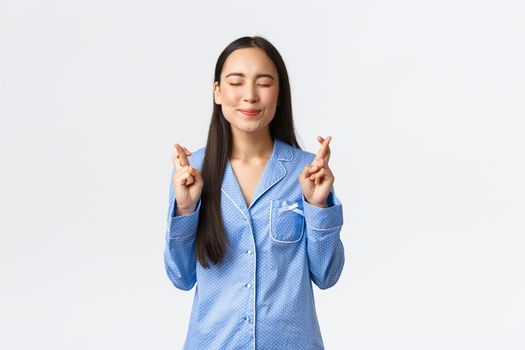 Hopeful happy asian girl in blue pajamas smiling with closed eyes, cross fingers good luck, making wish, praying or pleading for dream come true, anticipating positive news over white background.