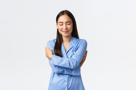 Happy dreamy tender asian girl in blue pajamas, close eyes and smiling as daydreaming, hugging herself, embracing own body in jammies, standing white background relaxed, daydreaming.