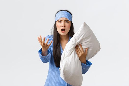 Angry bothered asian girl with insomnia, wearing sleeping mask and pajama, looking pissed-off as holding pillow and shaking hand furious, complaining on noise at night, white background.