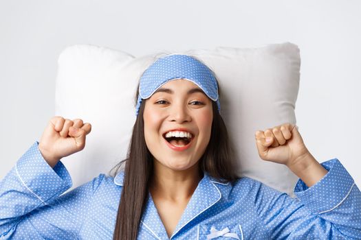 Close-up of enthusiastic asian girl in blue pajamas and sleeping mask, stretching hands up delighted after good night sleep, take-off eyemask in morning, lying in bed on pillow and smiling happy.