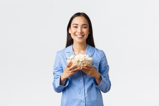 Home leisure, sleepover and slumber party concept. Smiling pleased asian girl enjoying weekends in bed with popcorn, eating and watching movies in pajamas, standing white background.