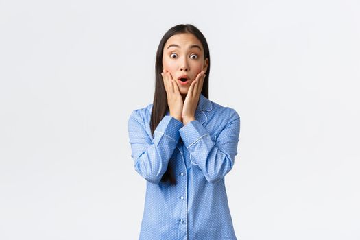 Surprised and astounded beautiful asian girl react to wonderful unexpected news, looking amazed with hands over face and opened mouth, standing in pajamas over white background.