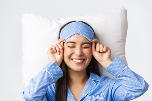 Close-up of happy beautiful asian girl in sleeping mask and blue pajamas lying in bed, smiling upbeat, having perfect mood as going bed and getting ready sleep, standing white background.