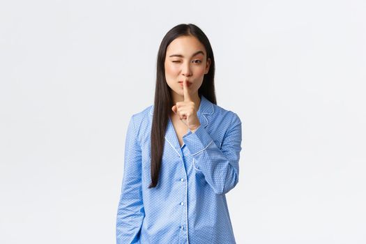 Attractive asian girl in blue pajamas hiding something, winking cunning and shushing, say shhh as asking keep quiet, showing silence gesture while standing over white background at sleepover.