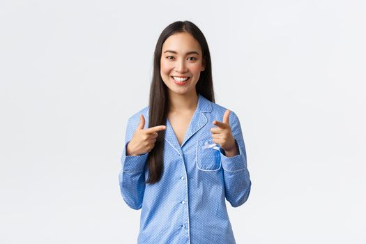 Cheerful cute asian girl smiling happy, standing in blue pajamas inviting girlfriends to sleepover party in jammies, pointing fingers at camera with confident joyful expression, white background.