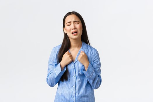 Miserable and sad asian girl in blue pajamas crying and complaining, feeling distressed, standing upset over white background, calling for help or grieving, posing over white background.