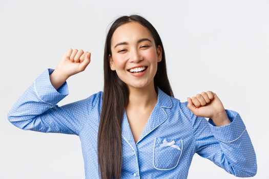 Close-up of cheerful smiling asian girl waking-up upbeat and stretching with happy face, had great night sleep, feeling energized starting morning with smile, white background.