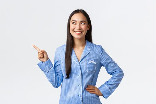 Satisfied happy asian woman in blue pajamas smiling broadly as pointing and looking at upper left corner. Pleased korean girl in jammies getting ready sleep, found great skincare product.