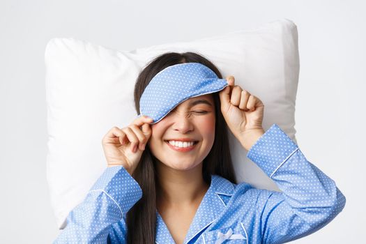Pleased happy smiling korean girl in blue pajamas and sleeping mask, had great day going bed in perfect mood, getting ready sleep, lying on pillow upbeat over white background.