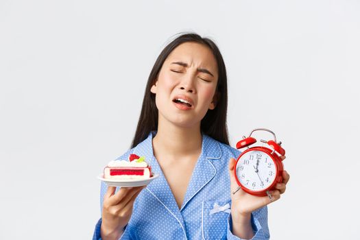 Close-up of displeased asian girl in blue pajama express regret as cant eat delicious piece of cake its too late at night, showing alarm clock and dessert, complaining cruel life, white background.