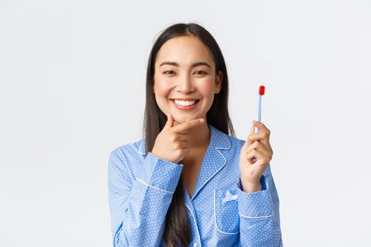 Close-up of attractive asian woman in blue pajama thinking while brushing her teeth with toothbrush, smiling and touching chin pondering great idea, white background. Copy space