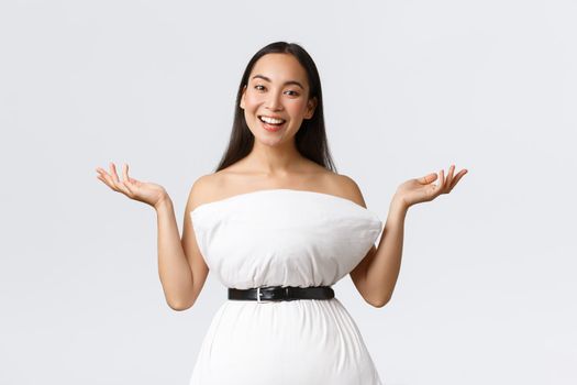 Gorgeous smiling and creative asian woman create dress from pillow cinching with belt around waste raising hands up and grinning, showing her new outfit, standing white background.