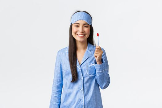 Hygiene, lifestyle and people at home concept. Cheerful smiling asian girl in pajamas and sleeping mask showing toothbrush and white perfect teeth, use whitening toothpaste, white background.