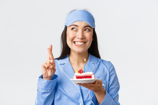 Celebration, lifestyle and holiday concept. Hopeful and dreamy smiling happy asian woman in sleeping mask and pajama, congratulated with birthday in bed, making wish before blowing candle on cake.