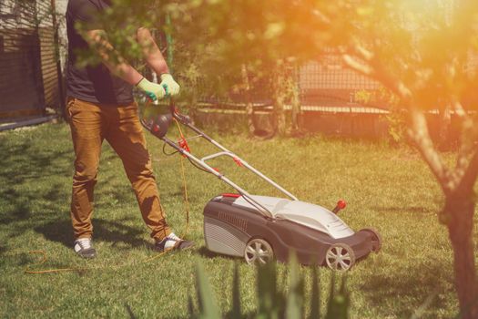 Young guy worker in casual clothes and gloves is mowing green grass with professional lawn mower on a yard. Gardening care equipment and services. Sunny day