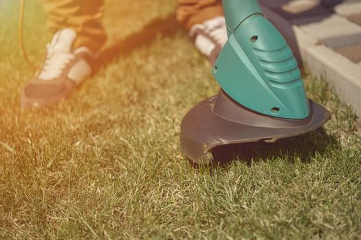 Man in sneakers and pants is mowing green grass with handheld electric lawn mower on his backyard. Gardening care equipment and services. Sunny day. Close up