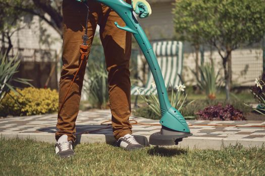 Guy in casual clothes and colorful gloves is cutting green grass with professional electrical mini lawn mower in garden of his country house. Deck chair, trees and flowers. Sunny day. Close up