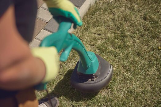 Professional gardener in sneakers and colorful gloves is mowing green grass with handheld modern lawn mower in courtyard, near a tiled path. Gardening care equipment. Sunny day. Close up, top view