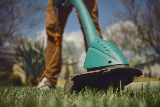 Male in casual outfit is mowing green grass with handheld electric lawn mower on his backyard. Gardening care tools and services. Sunny day. Close up