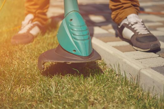 Male in sneakers and pants is cutting green grass with handheld modern lawn mower in his garden. Gardening care equipment and services. Sunny day. Close up