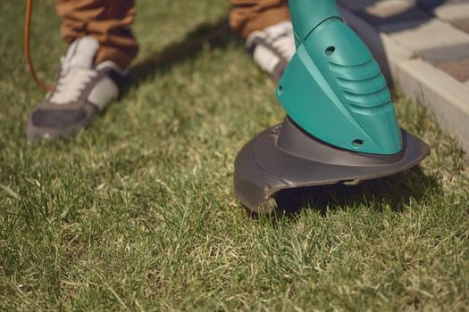 Man in sneakers and pants is mowing green grass with handheld electric lawn mower on his backyard. Gardening care equipment and services. Sunny day. Close up